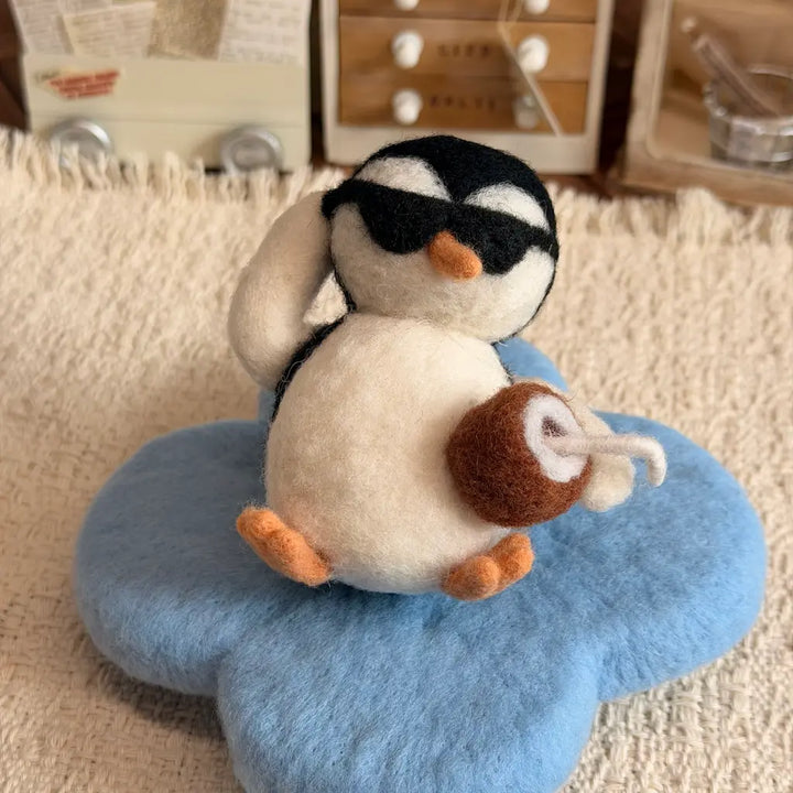 A small felt penguin plush wearing black sunglasses and holding a brown coconut drink, sitting on a blue flower-shaped cushion on a beige textured rug.