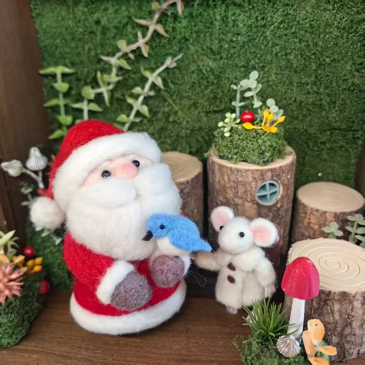 A festive miniature felt decoration set featuring a Santa Claus figure in red and white holding a blue bird, next to a small white mouse in a cozy woodland scene with faux moss, wood logs, a red mushroom, and greenery.