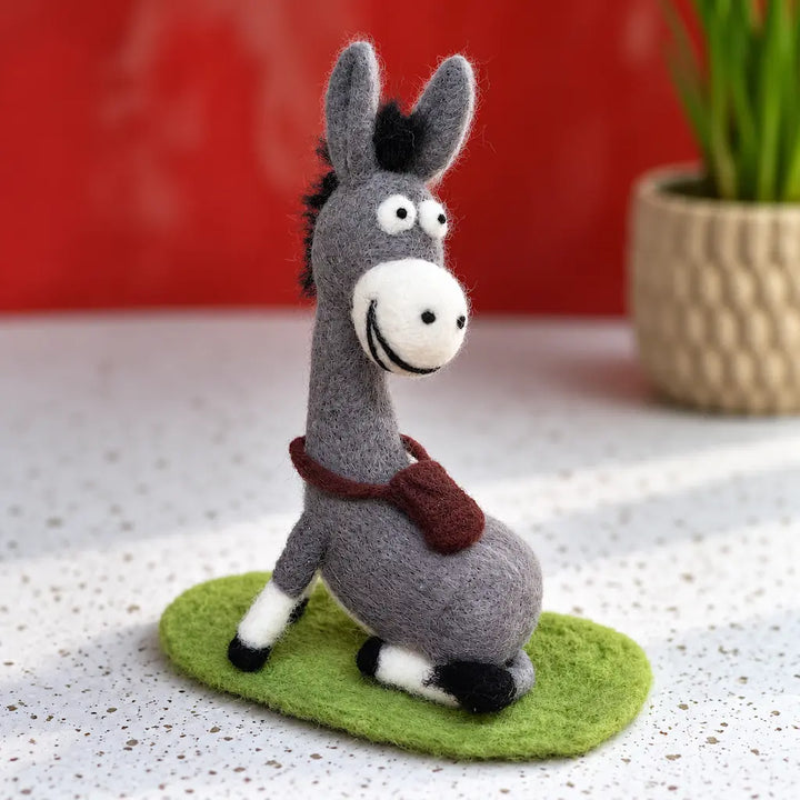 Front angled view of a seated gray wool felt donkey showing its cream muzzle, tall ears, brown satchel, and green mat.