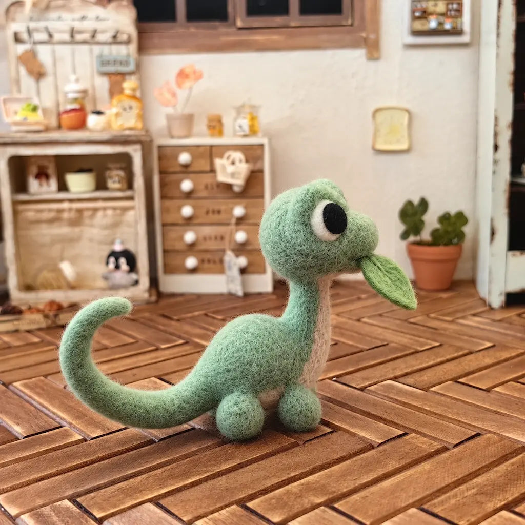Side view of a mint green wool felt dinosaur on a wooden tabletop, showing its leaf detail and curled tail.