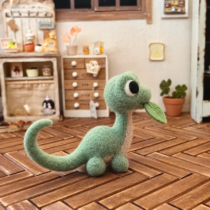 Side view of a mint green wool felt dinosaur on a wooden tabletop, showing its leaf detail and curled tail.