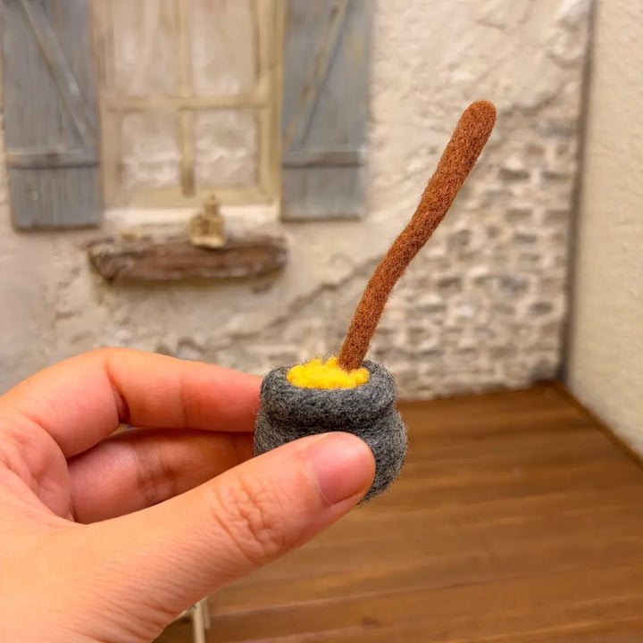 Close-up of a handmade charcoal-gray wool felt cauldron filled with yellow felt potion and a long brown stirring stick, held in a hand.