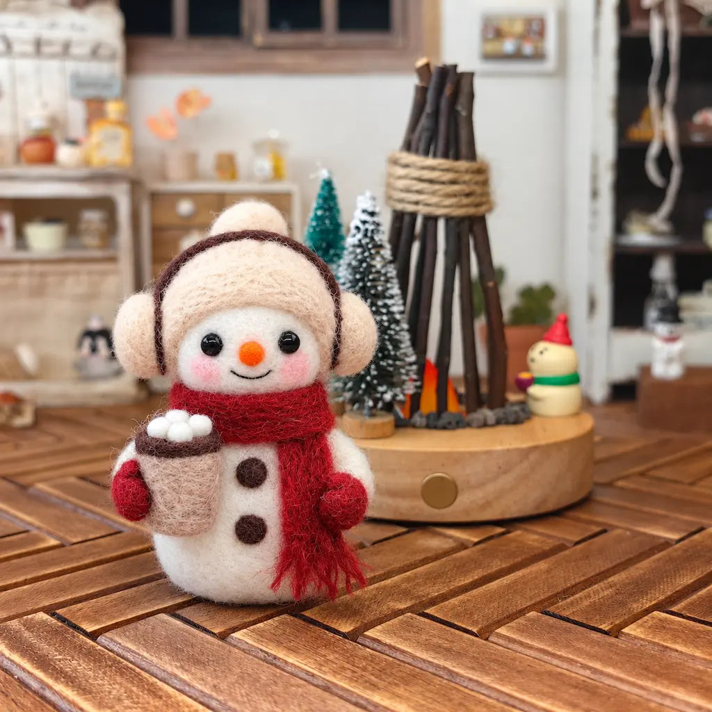 Close-up view of a handmade felt snowman holding a hot cocoa cup with marshmallows, wearing a cream hat and red scarf, styled with Christmas decorations on a wooden tabletop.