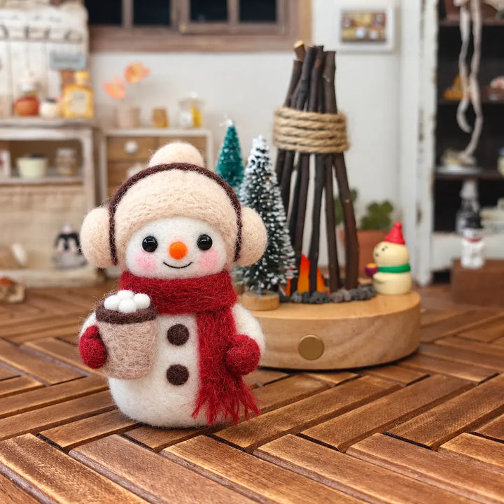 Close-up view of a handmade felt snowman holding a hot cocoa cup with marshmallows, wearing a cream hat and red scarf, styled with Christmas decorations on a wooden tabletop.