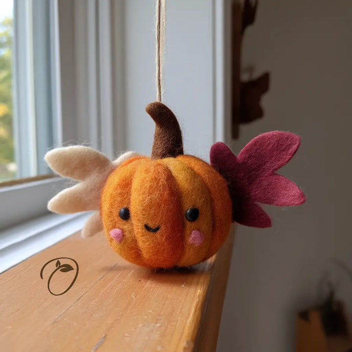 Front view of an orange wool felt pumpkin with ivory and cranberry leaf-shaped wings, pink cheeks, a curved brown stem, and a hanging cord.