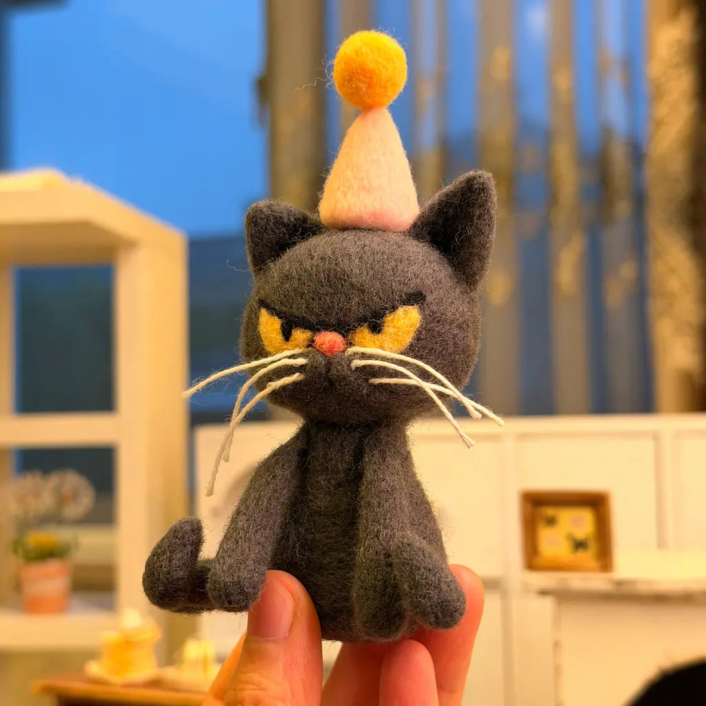 Three-quarter view of a handmade charcoal-gray wool felt birthday cat with an unimpressed expression, golden-yellow eyes, long whiskers, and a pink party hat.
