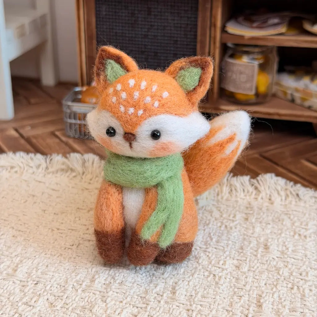 A small felted wool fox figurine wearing a light green scarf, sitting on a cream shag rug with a cozy wooden shelf and floor in the background.