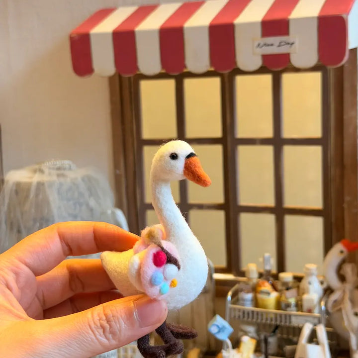 A hand holds a small felted wool swan figurine with a pink, white, and blue flower accessory. The background shows a miniature shop window with a red and white striped awning.