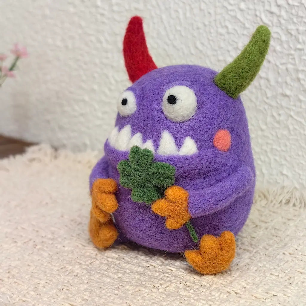 Handmade Clover Monster wool felt figurine with a round purple body, mismatched red and green horns, orange hands and feet, holding a green four-leaf clover.