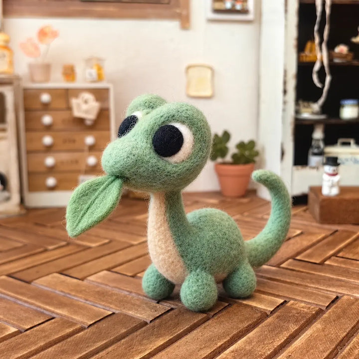 Angled front view of a wool felt dinosaur with oversized eyes, a cream belly, and a green leaf at its mouth.