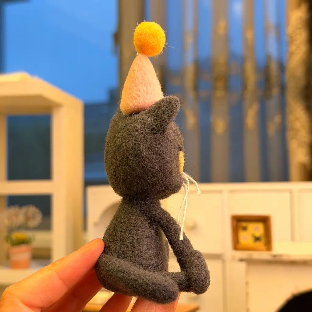 Side profile of a needle-felted gray birthday cat showing its pointed ears, elongated front paws, curled tail, and pink cone-shaped party hat with a yellow pom-pom.