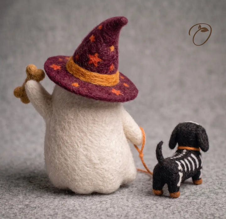 Handmade needlepoint figurine of a white ghost wearing a purple witch hat with orange stars and a yellow band, holding a small black-and-white skeleton dog on a gray textured background.