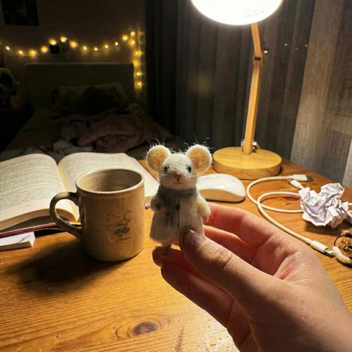Hand holding a small plush mouse toy on a desk with a mug, book, and lamp in the background.