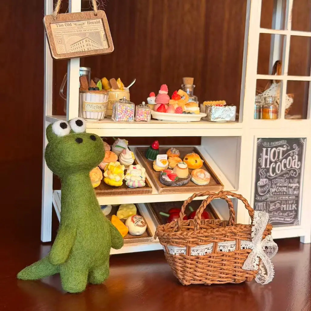 Green plush toy frog standing next to a miniature bakery display with a basket on a wooden floor.