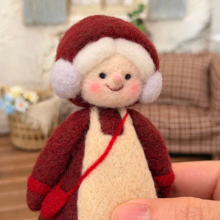 Hand-held front view of a handmade Mrs. Claus wool felt figurine showing her burgundy bonnet, soft white hair, cream apron, and red crossbody bag.