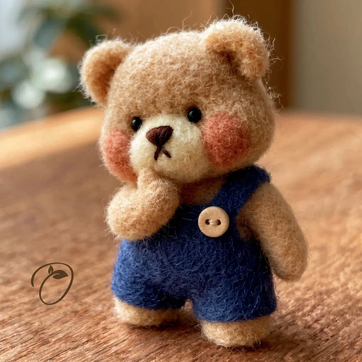 A small felt teddy bear figurine wearing blue overalls with a wooden button, standing on a wooden surface.