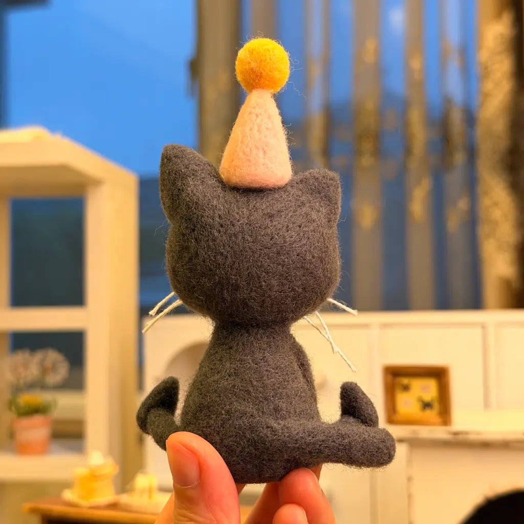 Back view of a handmade dark gray wool felt birthday cat with pointed ears, a curled tail, and a pale pink party hat topped with a yellow pom-pom.