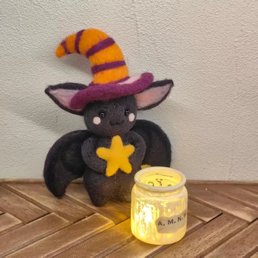 A small black plush bat toy wearing a pointed witch hat with orange and purple stripes and holding a yellow star, posed next to a glowing glass jar on a wooden surface against a textured wall.