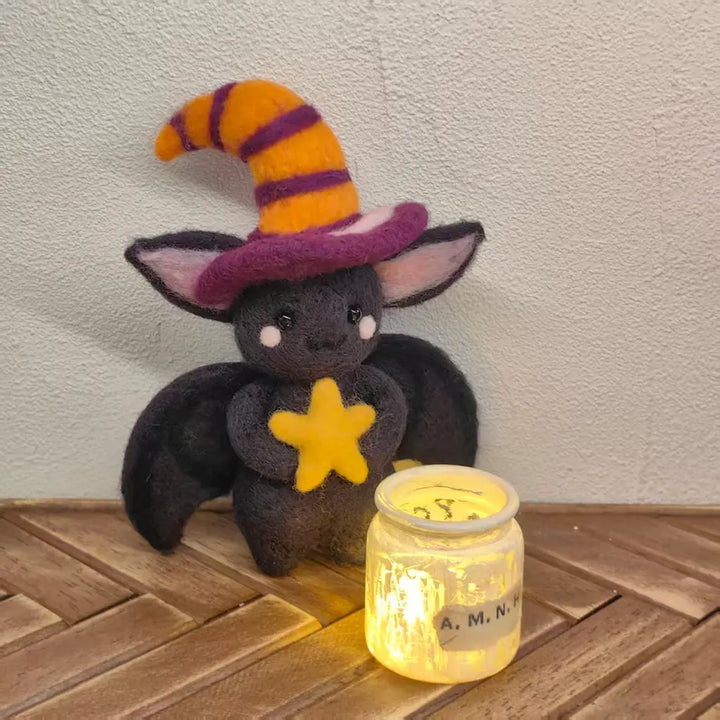 A small black plush bat toy wearing a pointed witch hat with orange and purple stripes and holding a yellow star, posed next to a glowing glass jar on a wooden surface against a textured wall.