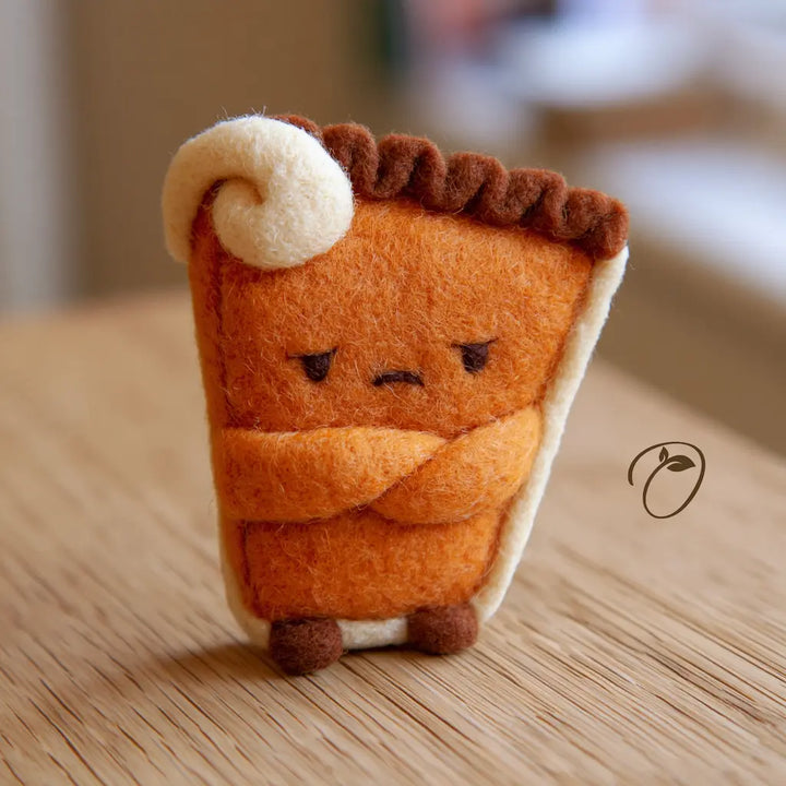 Front view of a wool felt pumpkin pie figurine with folded arms, a tiny frown, brown crust, and a cream-colored swirl.