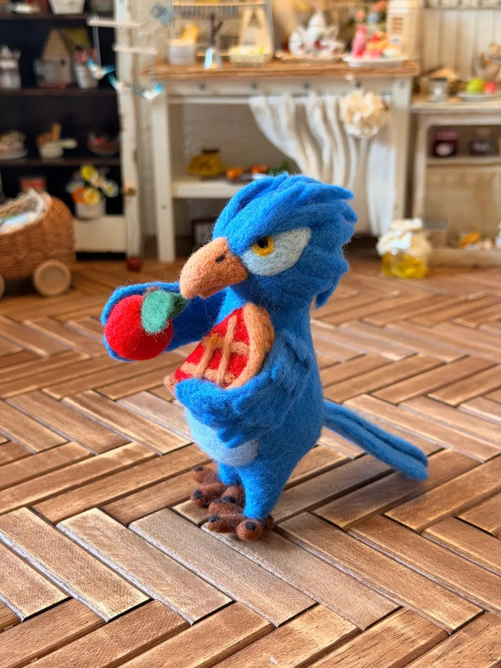 A blue felt parrot toy holding a red apple and a slice of pizza, standing on a wooden herringbone floor in a miniature room setting.
