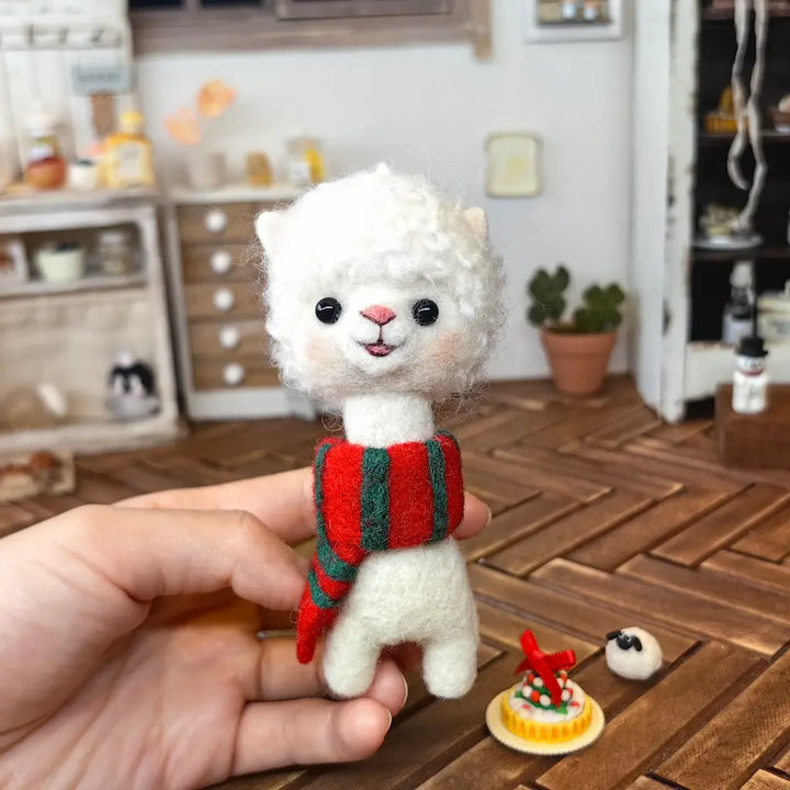 A small cream felt alpaca wearing a red-and-green scarf held in a hand above a wooden tabletop, with miniature holiday props nearby.