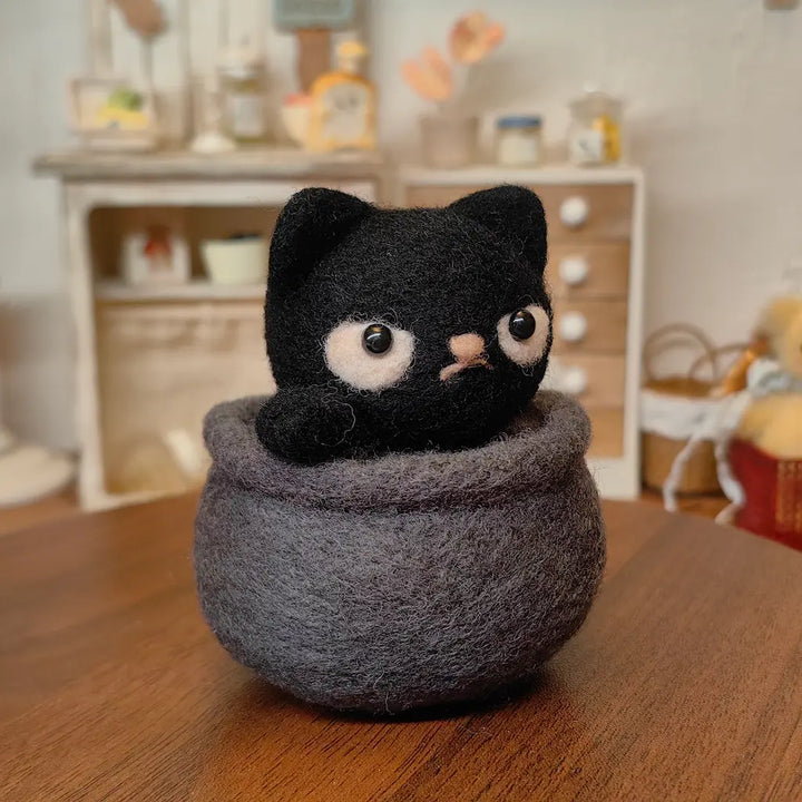 Grumpy black felt cat resting inside a handmade gray cauldron in a cozy indoor setting