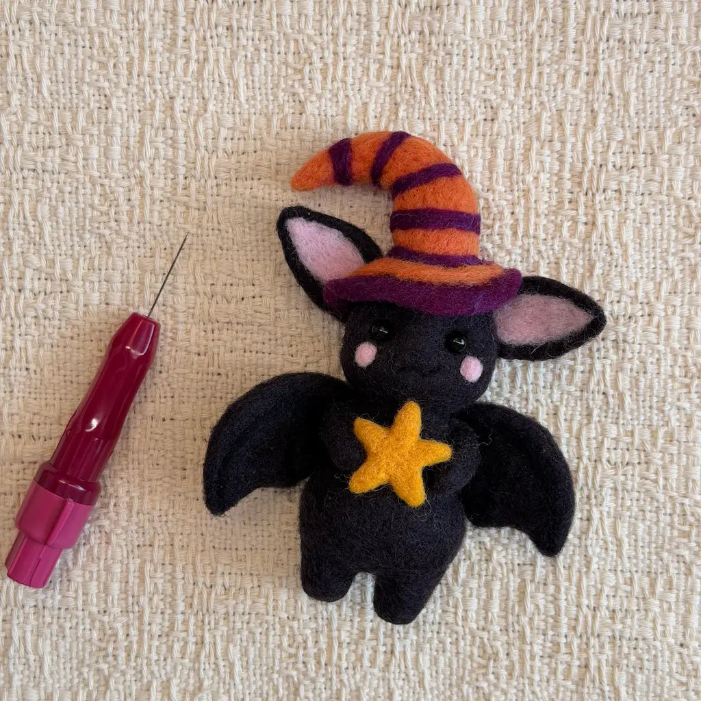 A small black felt bat plush toy wearing an orange and purple striped witch hat and holding a yellow star, placed on a beige textured fabric next to a pink needle tool.