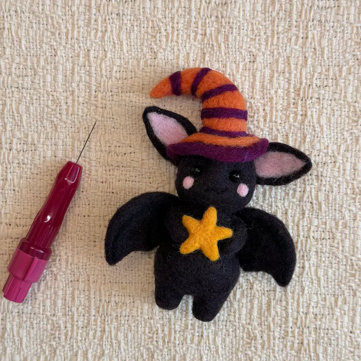 A small black felt bat plush toy wearing an orange and purple striped witch hat and holding a yellow star, placed on a beige textured fabric next to a pink needle tool.