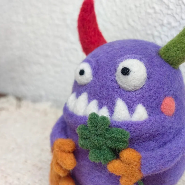 Close-up of the purple Clover Monster felt figurine showing its soft wool texture, mismatched horns, playful toothy grin and handmade green clover.