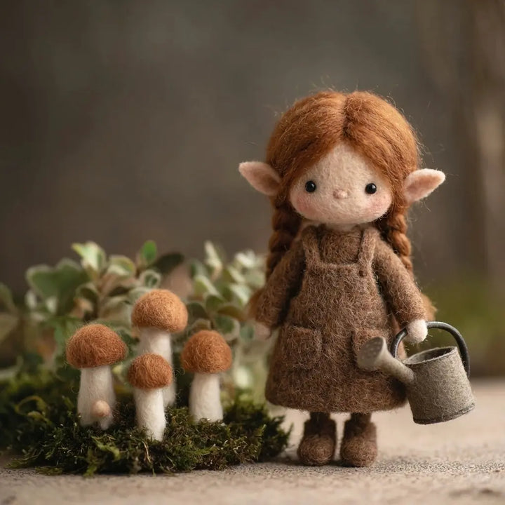 Felted fairy doll with brown hair and outfit standing next to mushrooms in a forest setting.