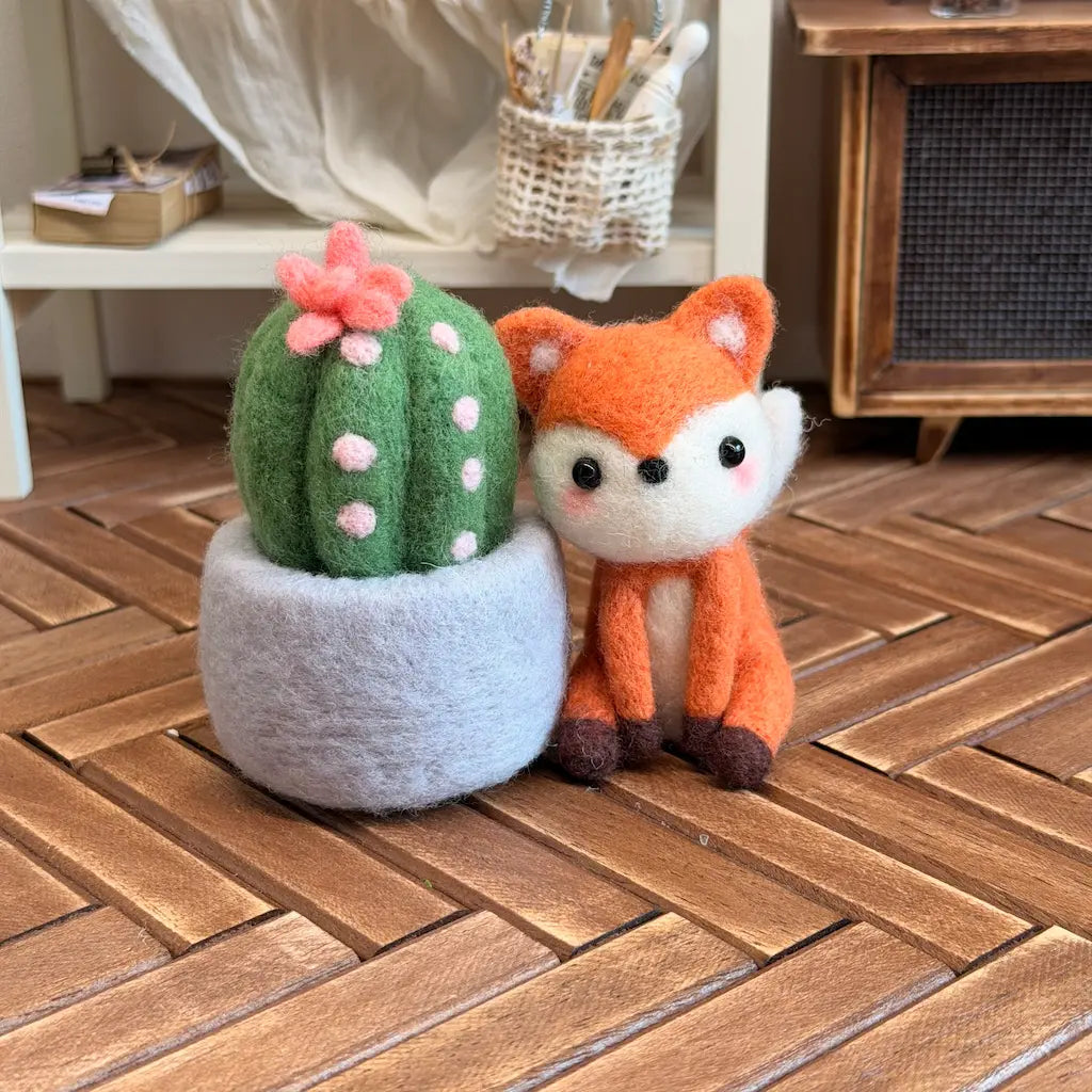 A felt needle art decoration set featuring a green cactus with pink spots and a pink flower in a gray pot, next to an orange fox with a white muzzle, displayed on a wooden herringbone floor.