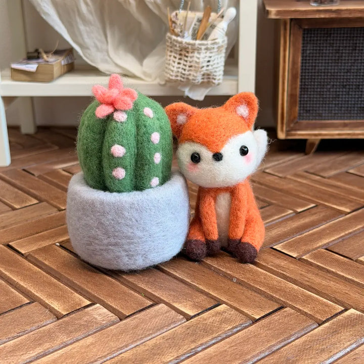 A felt needle art decoration set featuring a green cactus with pink spots and a pink flower in a gray pot, next to an orange fox with a white muzzle, displayed on a wooden herringbone floor.