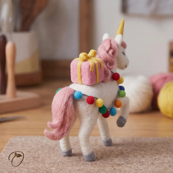 Left-facing felt unicorn with a curled pink mane, closed eyes, one front leg raised and a pink present on its back.