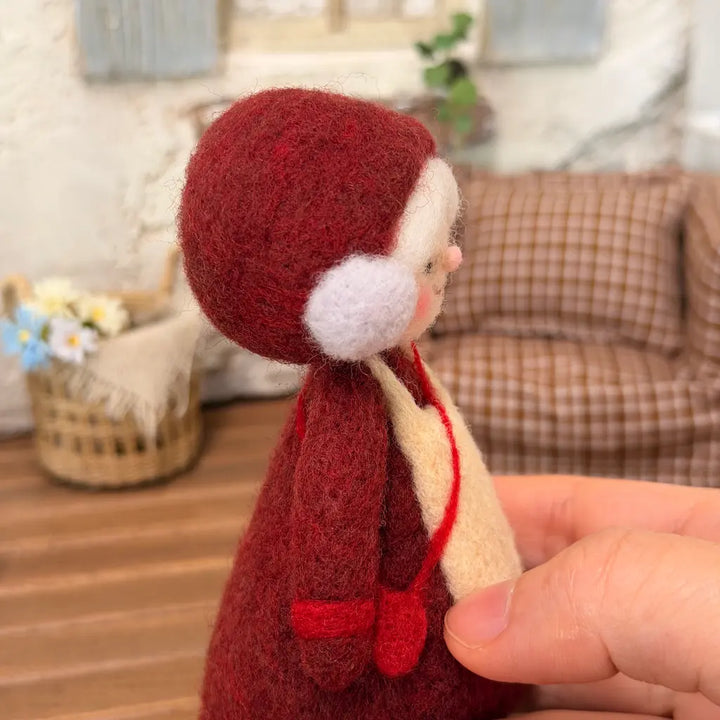 Hand-held side view of a needle-felted Mrs. Claus figurine highlighting her rounded burgundy bonnet, white wool curls, and textured burgundy dress.