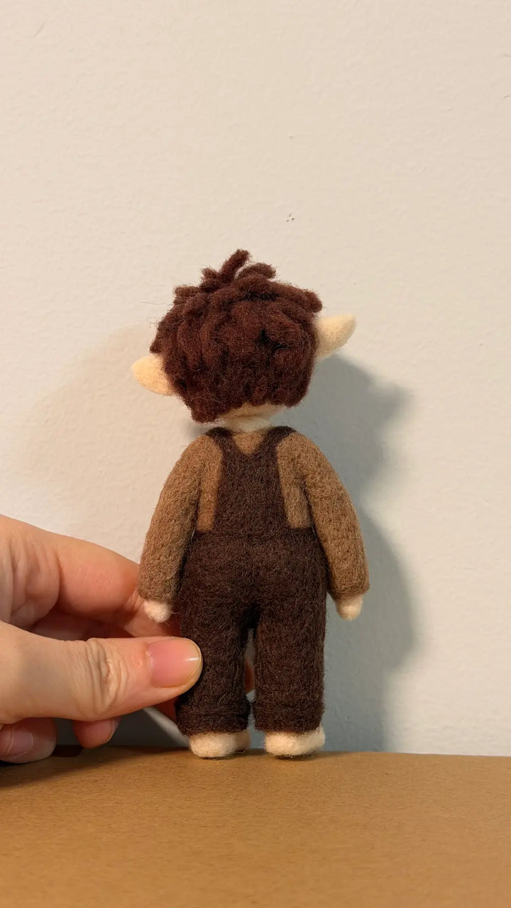 Small toy figure of a character with brown hair and overalls held by a hand against a plain background.