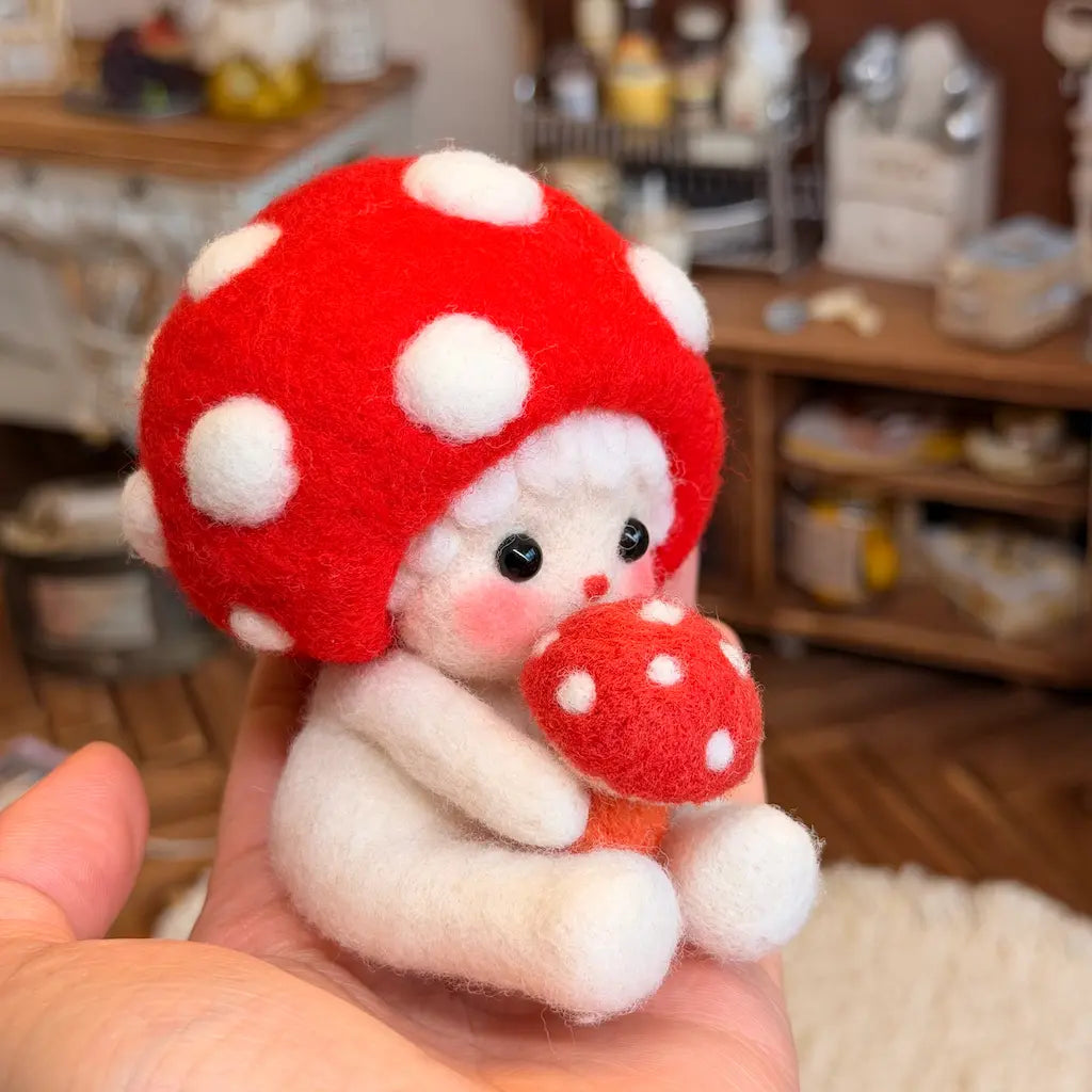 A small white felt doll with a large red mushroom cap and holding a smaller red mushroom, being held in a person&