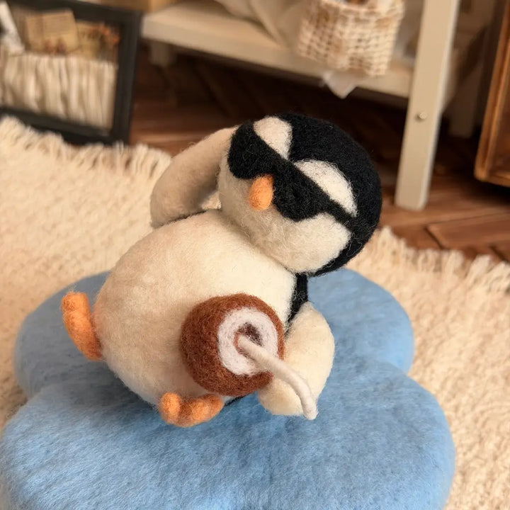 A felt penguin plush toy wearing black sunglasses and holding a brown drink, sitting on a blue cloud-shaped cushion in a cozy indoor setting.