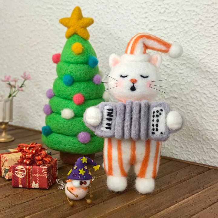 Handmade Accordion Cat wool felt figurine in orange-striped pajamas playing a gray accordion beside a colorful felt Christmas tree and wrapped gifts.