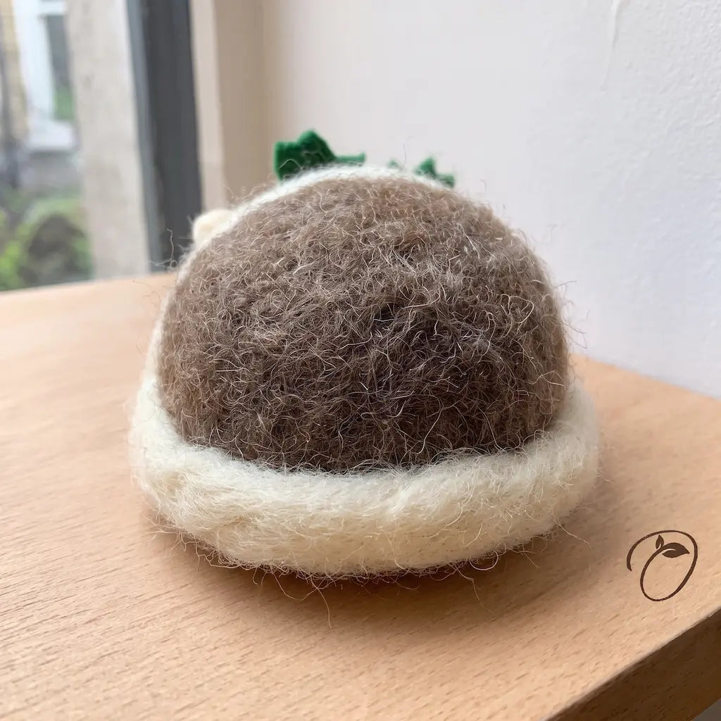 Back view of a brown felt hedgehog with cream trim around its rounded body, on a wooden surface beside a window.