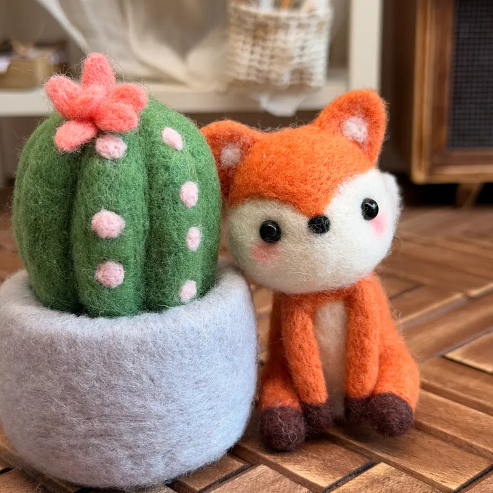 A felt needle art toy set featuring a green cactus with pink polka dots and a pink flower in a gray pot, next to an orange and white fox, displayed on a wooden surface.