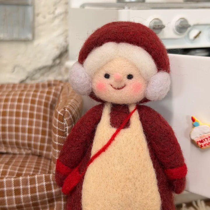 Close-up of a needle-felted Mrs. Claus figurine with a smiling face, rosy cheeks, fluffy white curls, burgundy bonnet, and cream apron.