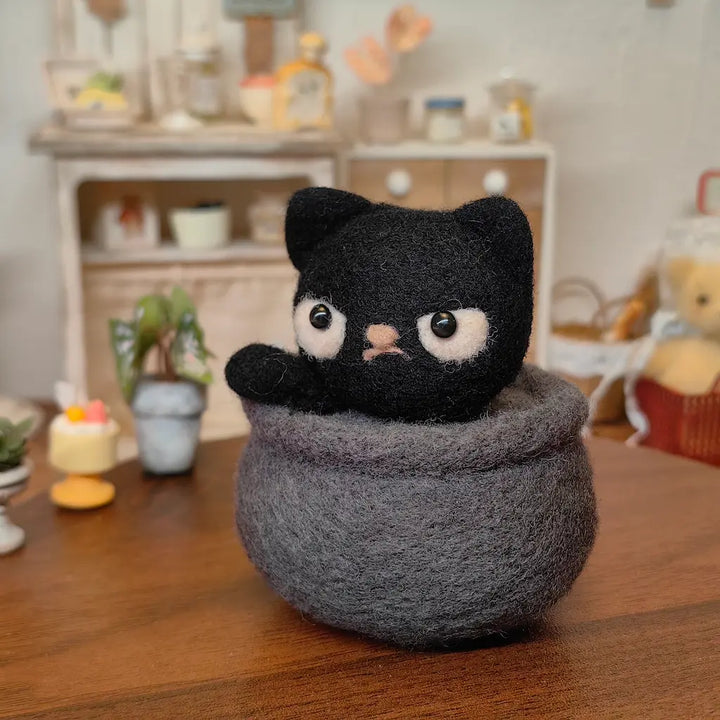 Black wool felt cat peeking from a charcoal-gray cauldron on a warm wooden table