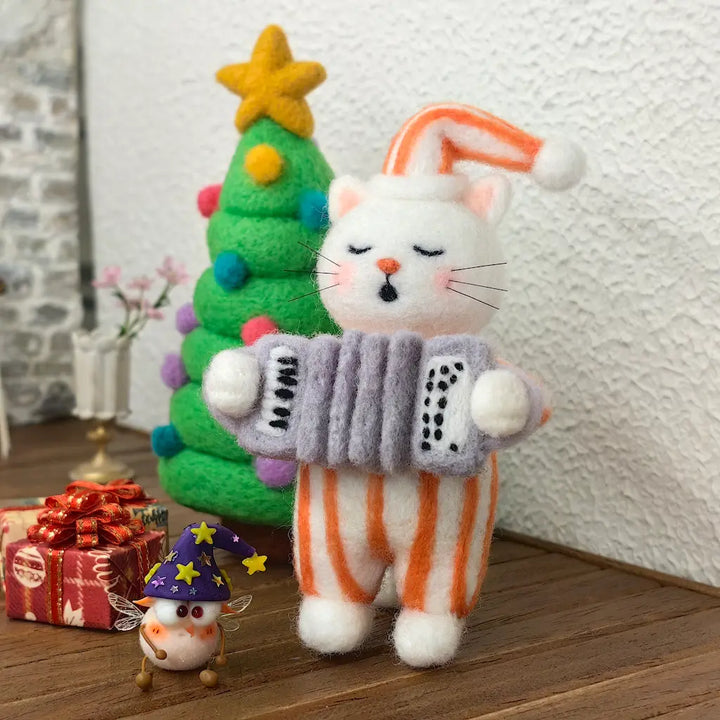 Close-up of a white needle-felted cat with closed eyes, an orange-striped sleeping cap and matching pajamas, holding a miniature gray accordion in a Christmas display.