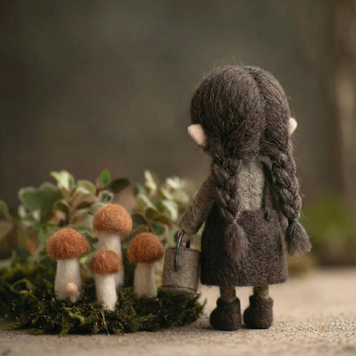 Felted doll with braided hair and a coat standing next to three felted mushrooms on a natural background.