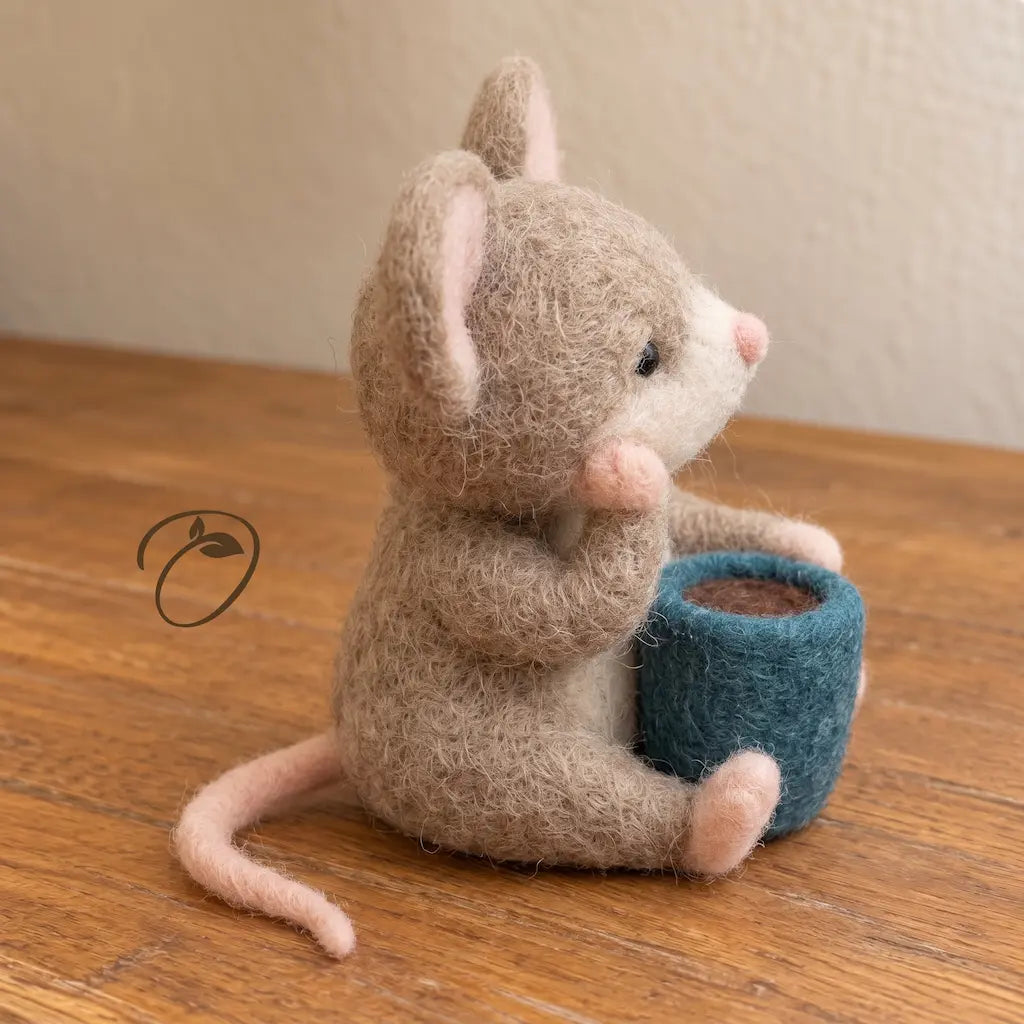 Side view of needle-felted Cozy Cup Mouse seated beside a blue mug