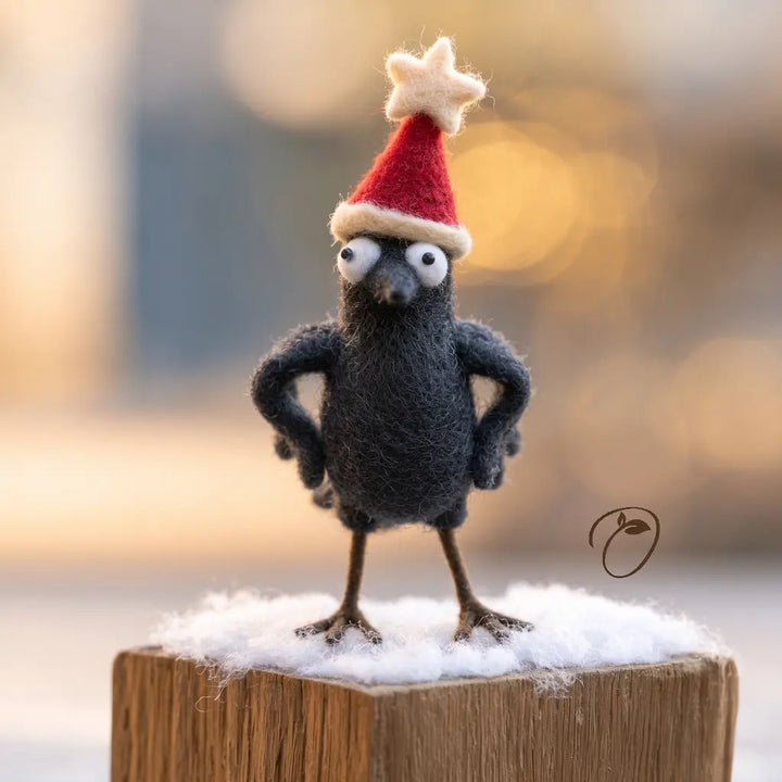 Front view of a black felt crow with wide white eyes, wings on its hips and a red star-topped Christmas hat.