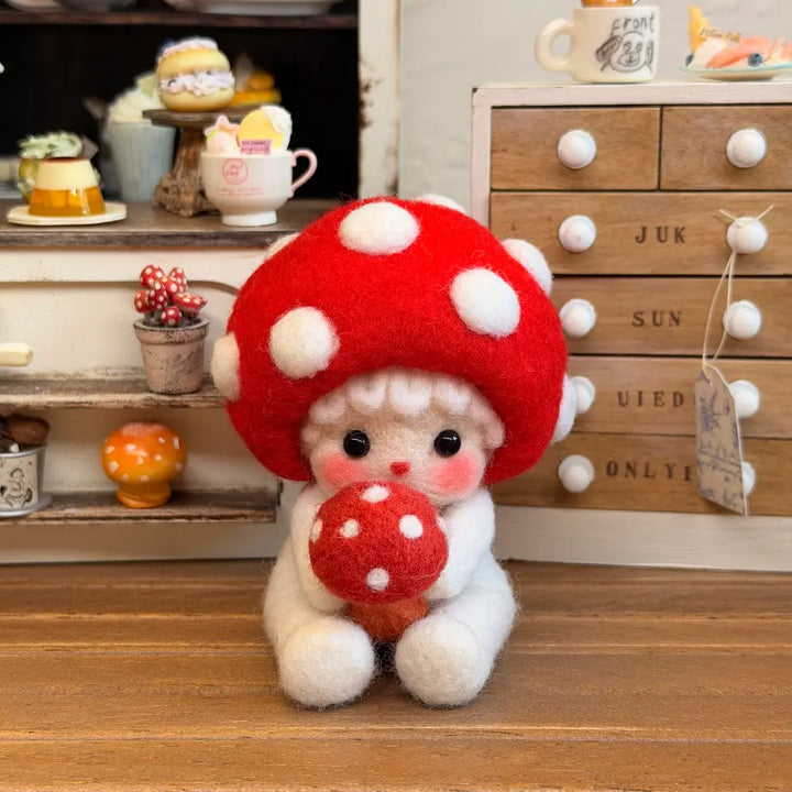 A small felted wool doll with a large red mushroom cap and white polka dots, sitting on a wooden surface and holding a matching mushroom. The doll is photographed in a miniature room setting with tiny furniture and accessories in the background.