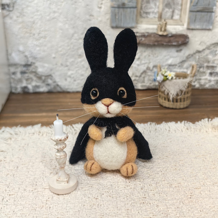 Handmade black masked bunny wool felt figurine with tall ears, a superhero cape and cream-colored belly, displayed beside a miniature candle on a cozy tabletop.