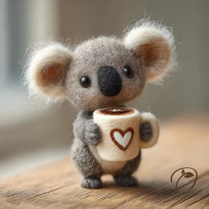 Felted koala toy holding a coffee cup with a heart design on a wooden surface.